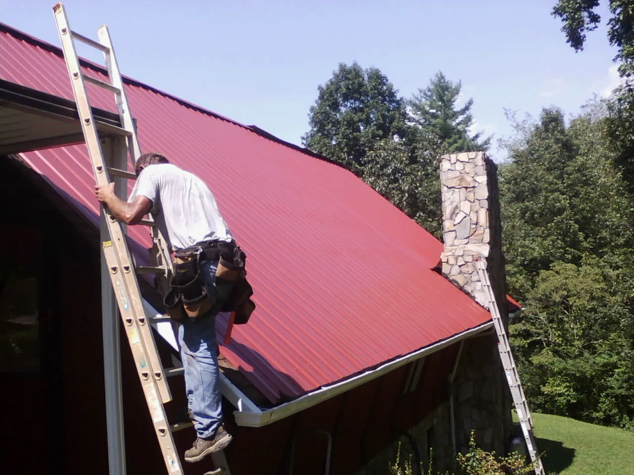 Roofer on a ladder beside a red metal roof and stone chimney — Soffit & Fascia Repair by Roofing Brothers in Sharpes
